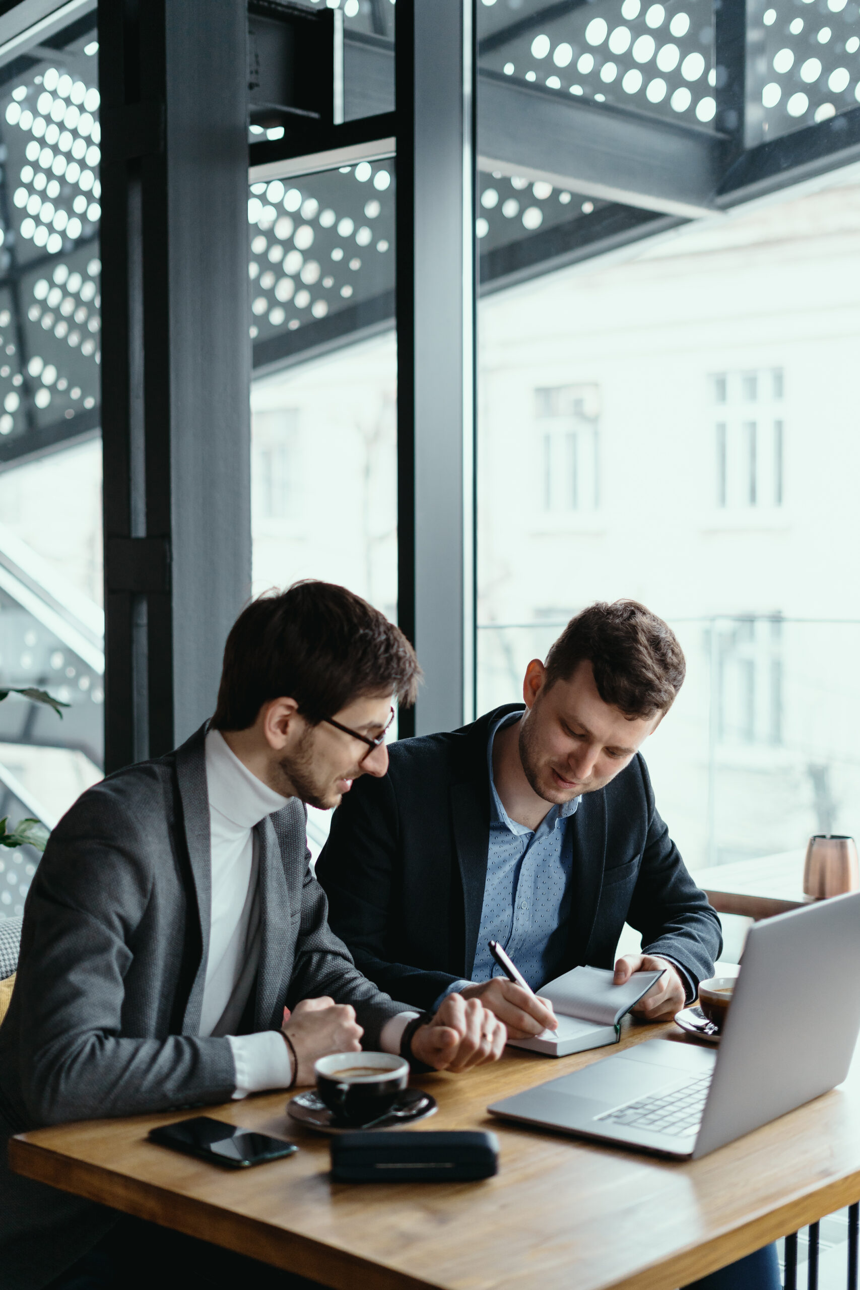 Two businessmen talking about new opportunities sitting with laptop at desk near large window, planning project, considering business offer, sharing ideas while drinking coffee together